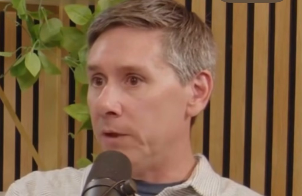 Mark a medium aged what man is speaking into a microphone in front of a wooden wall panel.