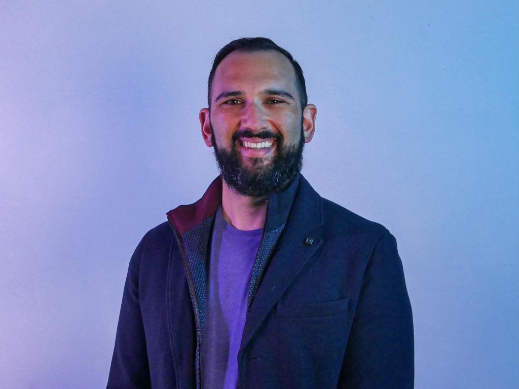 Jonathan, a white man with black hair and a black beard, is in a purple t-shirt and blue sports coat in front of a purple background.