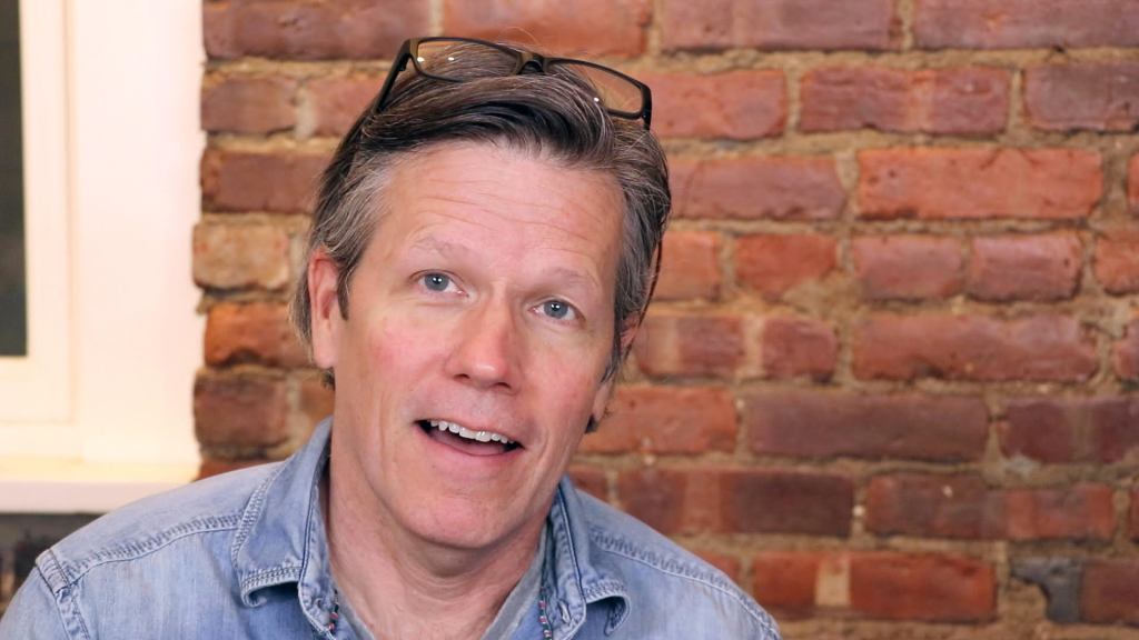 Mark, an older white man, has glasses atop his salt and peper hair. He has an open mouthed smile in front of a brick wall.